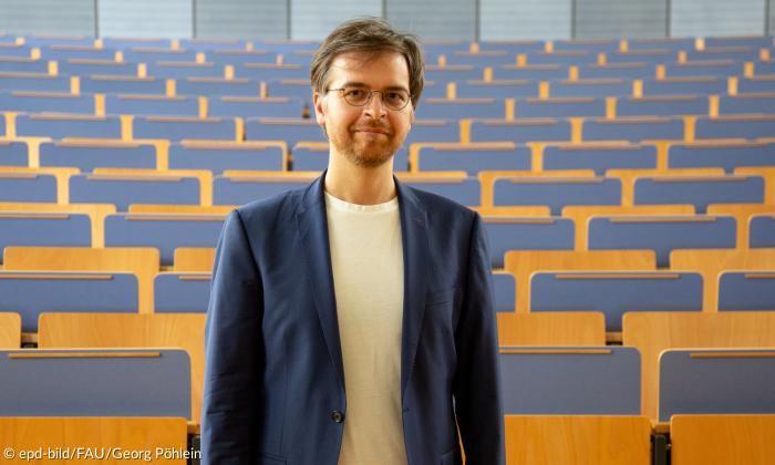 Ein Mann mit Jacket und Brille steht vor einem Hörsaal mit leeren Bankreihen