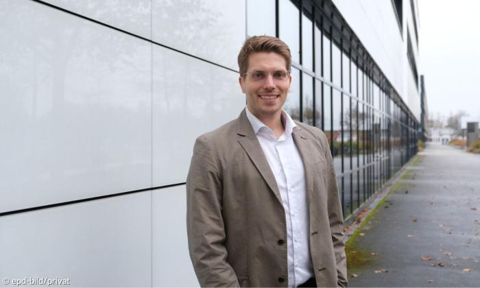 Ein Mann in Hemd und Jacke steht vor einer Hochschule