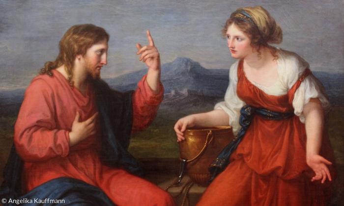 Ein Mann mit langen Haaren und eine Frau im roten Kleid unterhalten sich an einem Brunnen