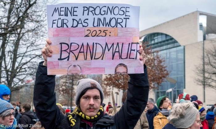 Ein Mann mit Mütze hält ein Schild hoch, auf dem steht: "Meine Prognose für das Unwort 2025: Brandmauer". Auch die jeweilige Stirn von Friedrich Merz und Alice Weidel ist zu sehen.