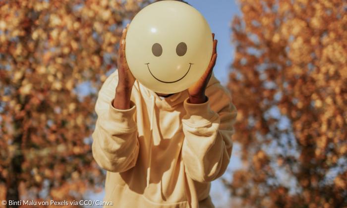 Ein Mensch hält einen Luftballon mit lachendem Smiley vor das Gesicht