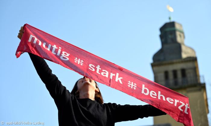 Eine Frau hält ein Banner hoch mit der Aufschrift "mutig, stark, beherzt"