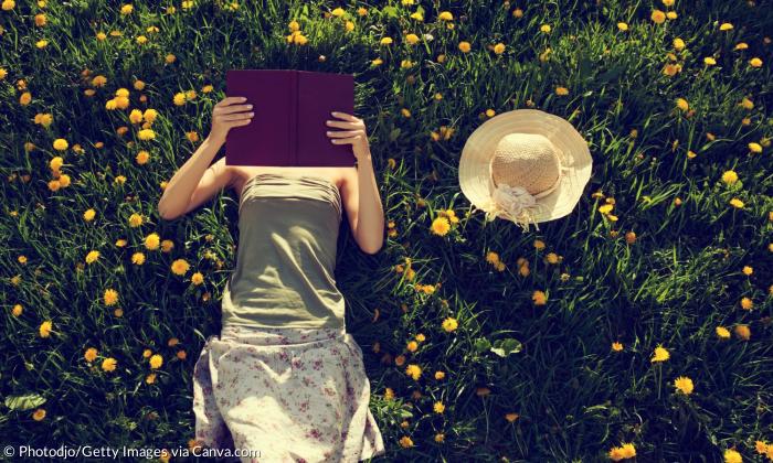 Eine Frau liegt auf einer Wiese, ein Buch vor dem Gesicht, neben ihr liegt ein Strohhut