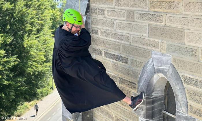 Ein Mann im schwarzen Talar mit einem grünen Helm seilt sich von einem Kirchturm ab