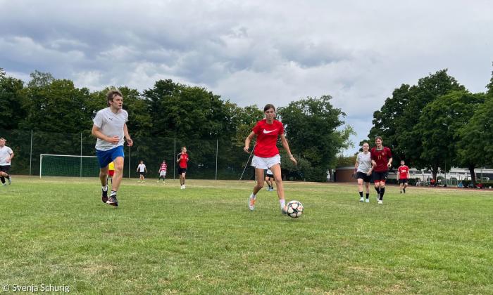 Menschen in Sportbekleidung auf einem Fußballplatz, eine Frau hat den Ball am Fuß