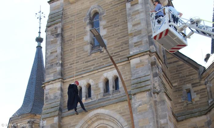 Ein Mann im Talar hängt am Seil an einer Kirche