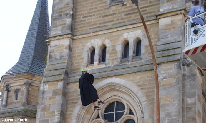 Ein Mann im Talar hängt am Seil an einer Kirche