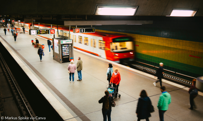 Menschen stehen vereinzelt an einer Ubahhaltestelle. Ein Zug fährt vorbei.