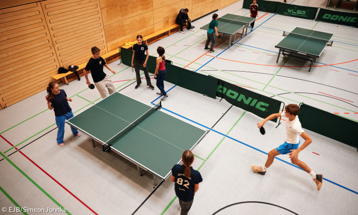 ugendliche stehen an einem Tischtennis-Tisch in der Turnhalle, der Ball fliegt gerade über das Netz. Im Hintergrund sind weitere Teilnehmer:innen bei anderen Aktivitäten zu sehen.