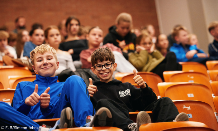 Zwei Kinder sitzen in einer Reihe und zeigen mit einem Daumen nach oben.