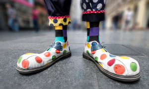 weiße, viel zu große Clownsschuhe mit bunten Punkten drauf. Die Socken haben auch ein buntes Muster und die Hose besteht aus unterschiedlichem Stoff. 