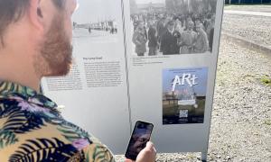 Ein Mann steht vor einer Tafel. Auf der Tafel ist ein Plakat zur Aktion ARt zu sehen. Der darauf abgebildete QR Code wird gerade von seinem Handy gescanned