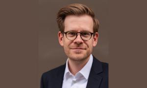 Ein Porträt von Andreas Braveny. Er trägt einen Anzug und eine Brille, lächelt in die Kamera und hat braune Haare.