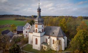 Das Gebäude der Markgrafenkirche Kirchgattendorf aus der Luft. Sie hat einen Kirchturm mit einer Uhr und besteht sonst aus einem nahezu viereckigen Hauptgebäude. Im Hintergrund sind Felder und herbstliche Bäume zu sehen.