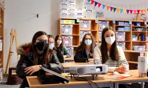 Kinder mit Masken sitzen an drei Tischen und blicken in die Kamera.