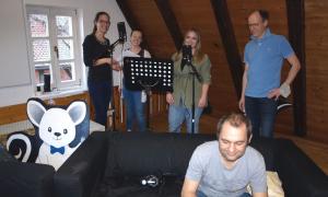 Vier Menschen stehen vor einem Notenständer und einem Mikrofon. Neben ihnen steht ein Pappaufsteller mit übergroßen Maus, die in die Kamera winkt. Vor den Menschen mit dem Mikrofon steht ein Sofa, auf dem ein Mann sitzt, der auf den Boden schaut. 