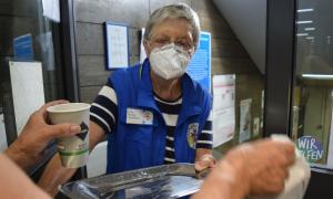Eine Frau in blauer Weste und gestreiftem Kurzarmshirt reicht einem Menschen ein Tablett und einen Kaffee.