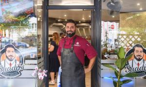 Ein Mann in einem roten, kurzärmligen T-Shirt und einer schwarzen Schütze steht vor dem Eingang eines Restaurants und lächelt in die Kamera.