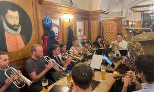 Zehn Personen sitzen in einem Wirtshaus um einen Tisch. Sie spielen Blechblasinstrumente. Trompeten und Tuba. 