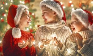 Drei Kinder haben Weihnachtsmützen auf dem Kopf und eine Lichterkette in der Hand und lachen sich an 