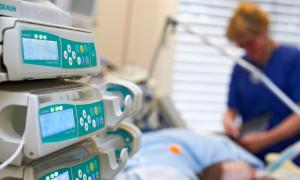 Links im Bildvordergrund sind medizintechnische Geräte. Verschwommen im Bildhintergrund sieht man eine Pflegerin am Bett eines Patienten