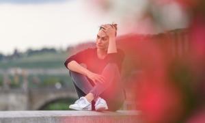 Eine junge Frau sitzt auf einer Mauer. Ihre Beine sind vor ihrem Körper über Kreuz aufgestellt. Mit ihrer linken Hand hält sie ihren Kopf.