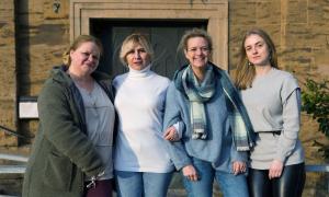 Vier blonde Frauen stehen nebeneinander vor einem Steinhaus. Sie schauen direkt in die Kamera und haben sich mit den Armen eingehakt.
