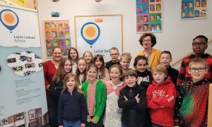 Eine Gruppe Grundschüler steht neben einem Banner mit der Aufschrift Luise Leikam Scvhule. Hinter den Kindern steht eine ältere Frau und an der Wand hängen Bilderrahmen mit bunten Popart Fotos von Luise Leikam.