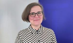 Eine Frau mit dunkler Brille und schwarz-weiß gestreifter Bluse lächelt mit geschlossenem Mund. Der Hintergrund ist auf der linken Seite hellgrau und auf der rechten Seite blau.