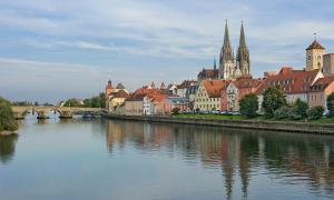 Im Vordergrund ist ein Fluss zu sehen. Am Ufer entlang ist die Regensburger ALtstadt und der Dom zu sehen, sowie eine steinerne Brücke, die über den Fluss führt.