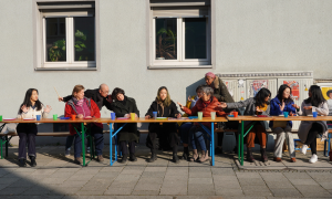 Mehrere Menschen unterschiedlicher Kulturen sitzen an einem langen Tisch, der auf der Straße steht. Sie diskutieren lebhaft miteinander und sitzen alle auf einer Seite des Tisches.