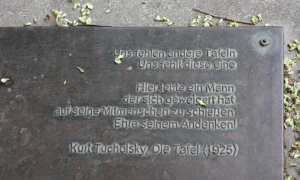 Eine Gedenktafel liegt auf dem Boden. Am oberen und rechten Bildrand liegen ein paar hellgrüne Blütenblätter herum. Auf der Tafel steht der Text "Die Tafel" von Kurt Tucholsky
