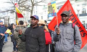Zwei Männer mit Fahnen und Schildern, dahinter mehr Demonstrant*innen