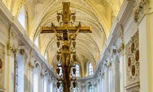 Bild des Hauptschiffes einer Kirche. Es strömt hell Sonnenlicht durch die großen Fenster. Im Vordergrund befindet sich das riesige goldene Kreuz mit einer Jesusstatue darauf.