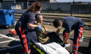 Man sieht, wie ein Mann von zwei Sanitäter*innen auf eine Trage gehievt wird. Er kneift schmerzerfüllt die Augen zusammen. 