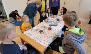 an einem Tisch sitzen Kleinkinder, die gerade Muffins essen. Eine ältere Dame beugt sich über den Tisch und spricht mit den Kindern. 
