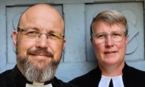 Ein Mann und eine Frau blicken frontal in die Kamera. Ihre  Man sieht nur ihren Kopf und ihren Schulterbereich. Sie wirken fröhlich, da sie die Mundwinkel leicht angewinkelt haben. Beide tragen einen Talar.