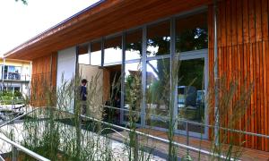 man sieht einen hölzernen Pavillon mit großer Glasfront; ein Mann tritt gerade durch eine Tür 