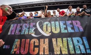 Ein großes Plakat mit der Aufschrift "Here & queer - auch wir!" 