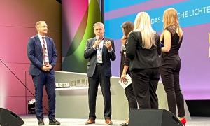 Ein Mann steht von vier Menschen umrundet auf einer Bühne; alle sind seriös gekleidet. Der eine Mann hat in der Hand ein Mikrofon
