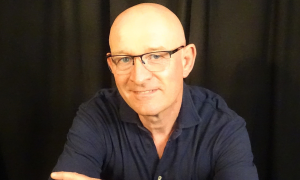 Portrait eines Mannes mit Glatze und Brille. Er trägt ein blaues Langarmhemd.