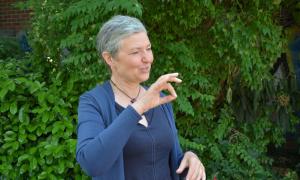 Seitenaufnahme einer Frau Mitte vierzig. Sie hat kurze graue Haare und gestikuliert mit ihrer rechten Hand, wobei Zeigefinger und Daumen ein Oval formen. 