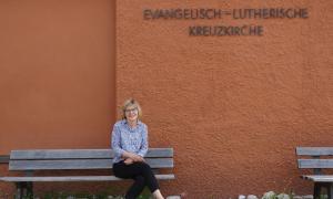 Eine Frau sitzt auf einer Bank vor einer rotbraunen Wand, auf der "Evangelisch-Lutherische Kreuzkirche" steht