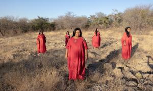 Fünf Frauen in roten Kleidern stehen in einer ausgetrockneten Graslandschaft.