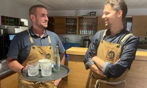 Zwei Männer mit Schürze, der eine hat ein Tablett mit Gläsern, einer Tasse und einer Flasche in der Hand, im Hintergrund eine Bar und Gläser