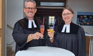 Ein Mann und eine Frau im Talar an einem Stehtisch mit Getränken in der Hand
