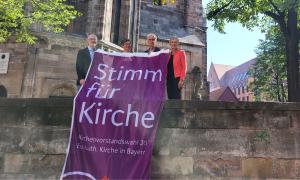 Bernhard Petry, Britta Müller, Jörg sichelstiel, Berthild Sachs mit dem Banner zu den Kirchenvorstandswahlen 2024 vor der Nürnberger Kirche St. Sebald. 