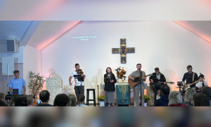 Mehrere Menschen mit Instrumenten stehen vor einem Kreuz auf einer Bühne
