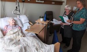 Zwei Frauen singen am Bett einer liegenden Seniorin