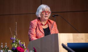 Eine Frau mit rotem Blazer und roter Brille spricht an einem Podium